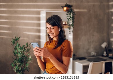 Young woman using smartphone while standing in a modern interior room at home. Communication, connection, mobile apps, technology, learning, chatting, lifestyle concept. Copy space. - Powered by Shutterstock