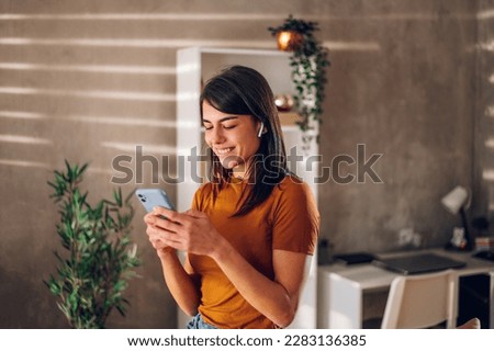 Young woman using smartphone while standing in a modern interior room at home. Communication, connection, mobile apps, technology, learning, chatting, lifestyle concept. Copy space.