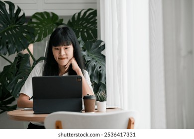 A young woman using a tablet at a home office desk, surrounded by green plants and natural light, creating a modern and relaxed workspace. - Powered by Shutterstock