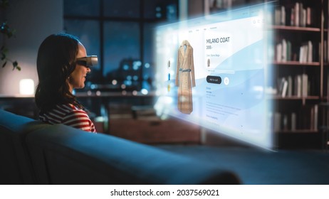 Young Woman Using Virtual Reality Headset At Home, Sitting on a Couch, Shopping Online via VR Clothing Store. Evening Resting at Apartment, Choosing New Look. Over the Shoulder