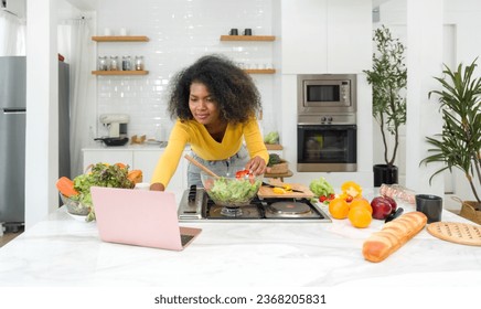 Young woman utilizing digital recipes from laptop computer. Cooking preparation in modern kitchen. - Powered by Shutterstock