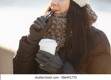 Young woman vapes & drinks hot cappuccino coffee from white paper mug outdoor in cold winter day.Vaper girl smokes vaporizer outdoor.Female model vaping outside - Powered by Shutterstock