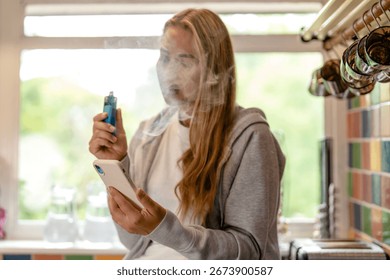 Young woman vaping while using smartphone in a bright kitchen during the afternoon