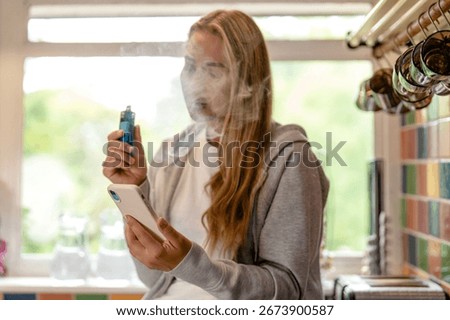 Young woman vaping while using smartphone in a bright kitchen during the afternoon