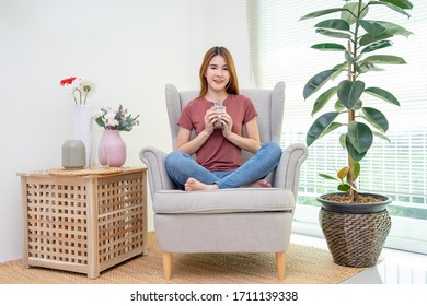 Young Woman Very Happy Putting Coin Into Jar For Saving Money On Sofa Near Window And Flower Vase, Indoors