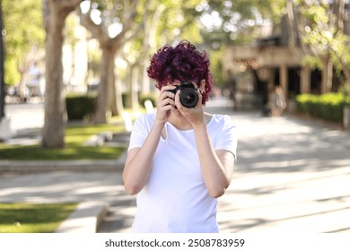 Young woman with vibrant purple curly hair taking a photo with a professional camera in a sunlit street surrounded by trees on a bright day, pointing to camera. Photography concept - Powered by Shutterstock