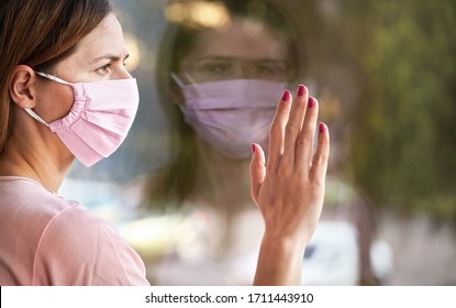 Young Woman In Virus Mask Looking Sad From The Window Behind Glass Pane, Touching It With Hand. Quarantine Or Stay At Home To Be Safe During Coronavirus Covid-19 Outbreak Concept