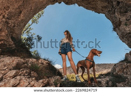 Young woman with vizsla dog standing in hole in rock. Female travel with pet in nature and climbing to mountain cave. Solo female slow travelling with pointer dog, hiking adventure in wild landscape.