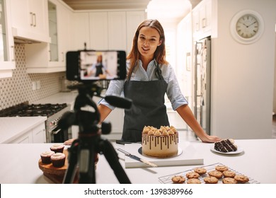 Young woman vlogger baking and recording video for food channel. Female pastry chef vlogging with her mobile phone mounted on a tripod in kitchen.