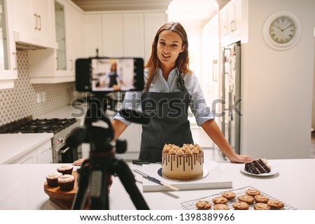 Young woman vlogger baking and recording video for food channel. Female pastry chef vlogging with her mobile phone mounted on a tripod in kitchen.