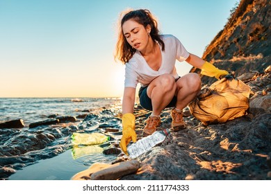 A young woman volunteer squats and picks up garbage on the ocean shore. Cleaning of the coastal zone. Sunset in the background. - Powered by Shutterstock