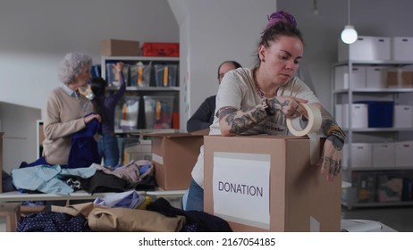 Young woman volunteer with tattoos use tape packing donation boxes in military warehouse. Group of people help army sorting and packing goods for humanitarian aid