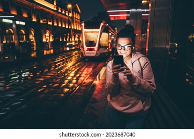 Young woman waiting for public transport inside modern transparent shelter at the night. 