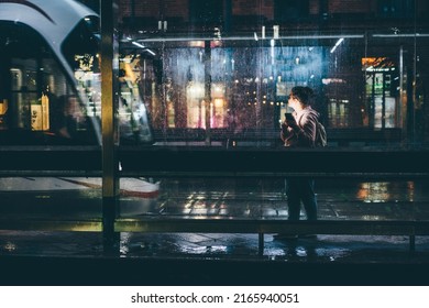 Young woman waiting for public transport inside modern transparent shelter at the night. 
