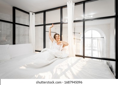 Young Woman Waking Up In The Beautiful White And Sunny Bedroom, Wide Interior View. Good Morning Concept