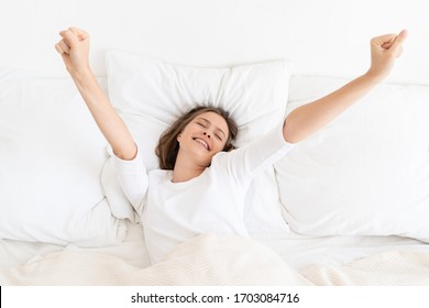 Young Woman Waking Up Late In Morning On Bed With White Sheets And Pillows, Dressed In T-shirt, Moving Arms Trying To Get Up, Eyes Closed, Smiling