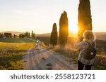 Young woman walking along the rural road with cypress trees at sunset. European tourist exploring the landscapes of Tuscany, Italy.