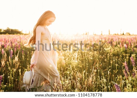 Young woman walking in lupine flower field with sunrise on the background. Warm orange sunbeam light