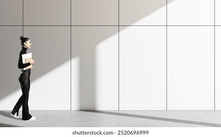 Young woman walking with a notepad in hand, in front of a modern tiled wall with sunlight casting shadows. Contemporary education concept