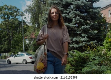 Young Woman Walking On City Street With A Reusable String Eco Bag Full Of Groceries. Young Girl Going Home After Shopping With String Mesh Bag With Vegetables. Conscious Consumption Concept