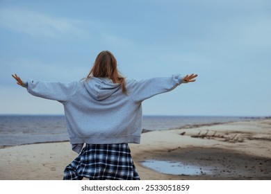 Young woman walking on a sandy beach with her arms outstretched, embracing the feeling of freedom and nature. Serene autumn day by the sea, symbolizing peace and liberation. - Powered by Shutterstock