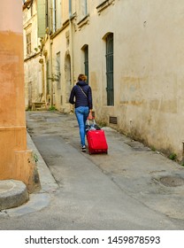 Young Woman, Walking, Pulling A Red Rolling Suitcase Down The Narrow Street Of The Old Town On Arles, France, April 2019