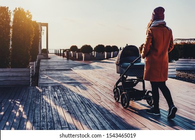 Young woman walking through city park with baby stroller. Warm autumn or spring weather for outdoor activity. Pretty urban architecture and landscape design. Sunset, clouds in sky, sunny day for walk