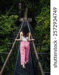 A young woman walks across a narrow suspension bridge in the heart of a lush, green forest. This serene and nature-focused scene perfectly captures the essence of travel and outdoor exploration.