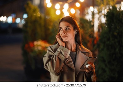 Young woman walks in the city at night, adjusting her wireless earphones and listening to music on her smartphone, surrounded by city lights and immersed in her own world - Powered by Shutterstock