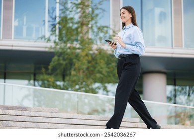 A Young Woman Walks Downstairs in Business Attire While Using a Smartphone on a Sunny Day Outside an Office Building - Powered by Shutterstock