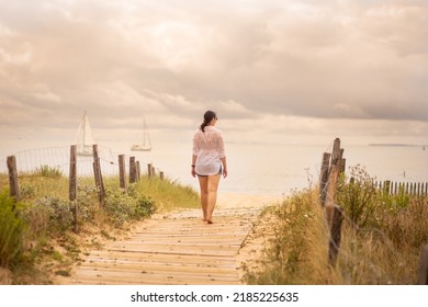 A Young Woman Walks On The Wooden Pontoon That Leads To The Beach. In The Background You Can See Two Boats With White Sails, Sailing In The Calm Water Of The Ocean. The Fence Forms A Path To The Beach