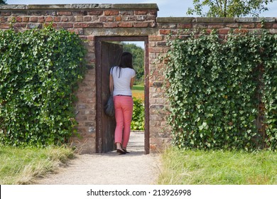 Young Woman Walks Through Wooden Doorway In An Ivy Clad Brick Wall. 