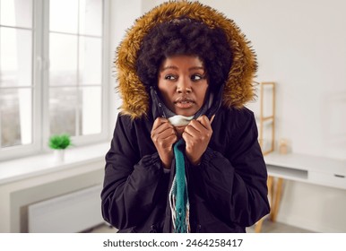 Young woman in warm clothes freezing inside the house during a cold winter. Shivering African American woman wearing a parka jacket and a scarf standing in a chilly living room at home - Powered by Shutterstock