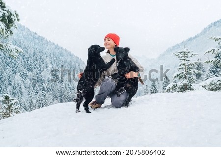 A young woman in warm clothes walking her 2 dogs in a picturesque snowy mountain outdoor. Female laughing and playing with pets and one dog licking an owner's cheek.Human and pets winter concept image
