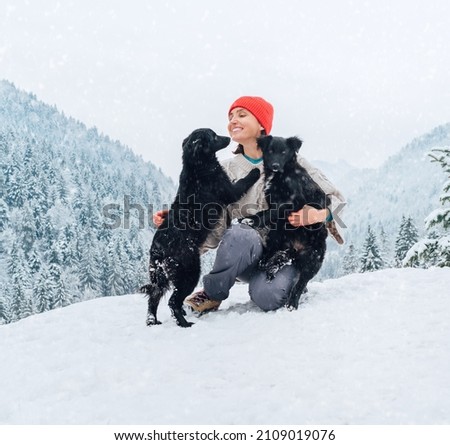 A young woman in warm clothes walking her 2 dogs in a picturesque snowy mountain outdoor. Female laughing and playing with pets and one dog licking an owner's cheek.Human and pets winter concept image