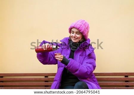 A young woman in warm outerwear pours hot tea from a thermos while sitting on a bench near the wall in a knitted pink hat and smiles cutely with happiness. Caucasian girl enjoying the moment