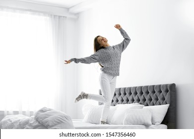 Young woman in warm sweater jumping on bed at home