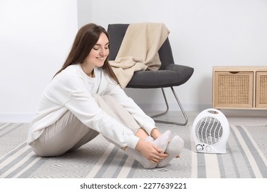 Mujer joven calentando pies cerca del calentador de ventilador eléctrico en casa