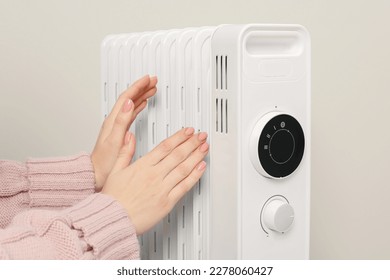 Young woman warming hands near modern electric heater on beige background, closeup