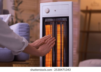 Young woman warming hands near electric heater in evening at home, closeup