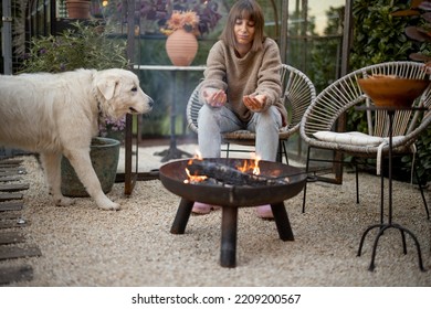 Young Woman Warms Her Hands, Spending Evening Time With Her Dog By The Fire At Cozy And Beautiful Backyard