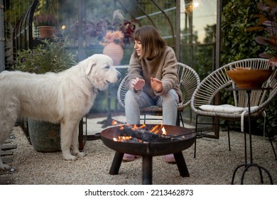 Young Woman Warms Her Hands, Spending Evening Time With Her Dog By The Fire At Cozy And Beautiful Backyard