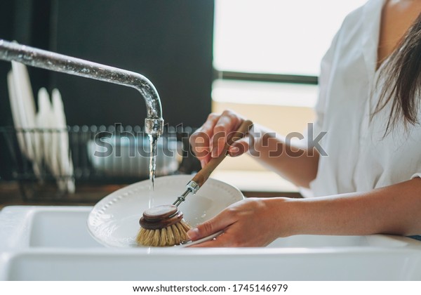 Young woman washes\
dishes with wooden brush with natural bristles at window in\
kitchen. Zero waste\
concept