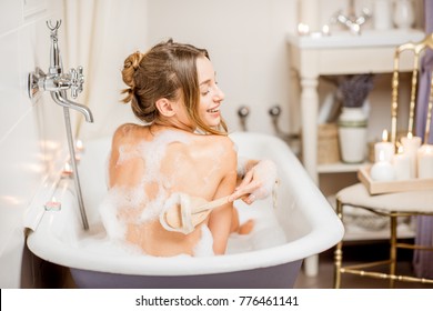 Young Woman Washing With Brush In The Beautiful Vintage Bath Full Of Foam In The Bathroom Decorated With Candles