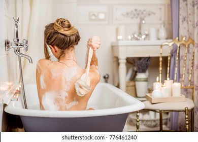 Young Woman Washing With Brush In The Beautiful Vintage Bath Full Of Foam In The Bathroom Decorated With Candles