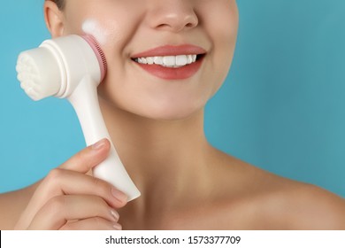 Young Woman Washing Face With Brush And Cleansing Foam On Light Blue Background, Closeup. Cosmetic Products