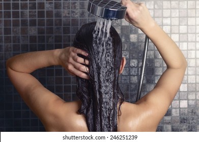 Молодая женщина мывает длинные волосы под душем стоя спиной к камере, смывая ее под струей воды с головой частично повернутой в сторону, над серыми мозаичными плитками