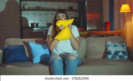 Young Woman Watching Horror Movie Sitting On Couch In Living Room At Night. Beautiful Scared Girl Hugging Pillow Watching Thriller Alone At Home