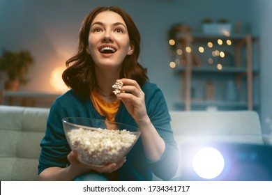 Young woman watching projector, TV, movies with popcorn in the evening. Girl spending time at home.