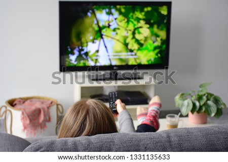 Young woman watching television with subtitles while sitting comfortably on sofa at home in living room. Nature, green, documentary, tv screen